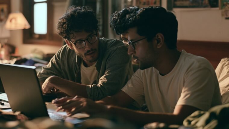 Two Indian students reviewing a video edit together on a laptop in a hostel room