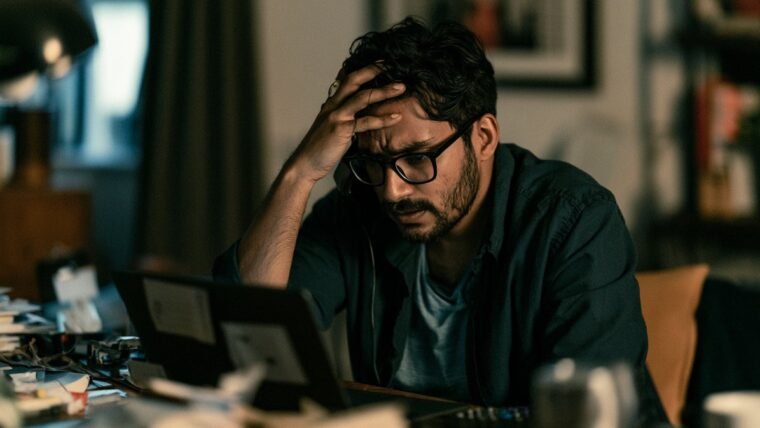 Man on hold on a phone call with one hand on his forehead at a desk